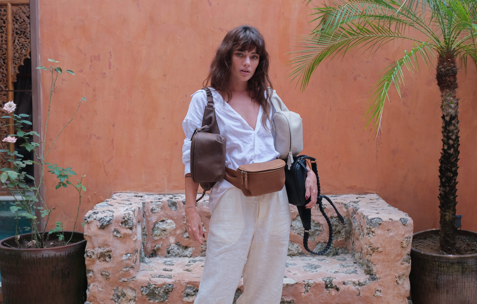 A woman stands in an outdoor courtyard wearing a brown leather crossbody bag and a matching brown leather belt bag, with a light gray duffel bag slung over her shoulder. The background features a textured terracotta wall, a stone bench, and potted plants.